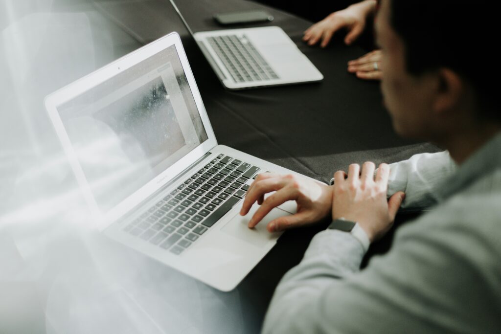 priscilla-du-preez-OEdkPaxYMXU-unsplash „Person arbeitet an einem Laptop während einer Besprechung, mit weiteren Laptops und Teilnehmern im Hintergrund.“