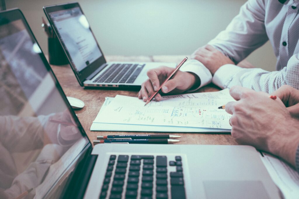 scott-graham-5fNmWej4tAA-unsplash „IT-Beratungssitzung: Zwei Fachkräfte arbeiten mit Laptops an einer technischen Strategie, während Notizen handschriftlich festgehalten werden.“